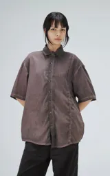 Camisa de corte oversize y mangas cortas, confeccionada en tela con efecto de teñido artesanal maltinto en tono gris. Presenta cuello clásico, cierre frontal con botones y costuras verticales decorativas en el frente.