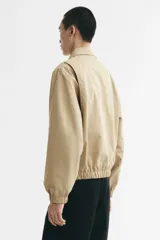 Campera tipo bomber de sarga de algodón en color beige, con cuello inglés, cierre frontal metálico y bolsillos diagonales ribeteados. Presenta terminaciones con elástico en los puños y el ruedo.
