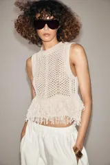 Chaleco de punto calado color crudo, de cuello redondo y sin mangas. El bajo está terminado con flecos.