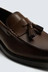 Mocasín de vestir color marrón oscuro, con parte superior lisa, detalle de antifaz con borlas en el empeine y horma redonda. Suela de goma a tono.