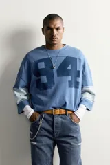 Camiseta boxy fit de color azul con efecto lavado vintage, cuello redondo y mangas por debajo del codo. Presenta un gran número '94' estampado en el frente y franjas horizontales en las mangas.