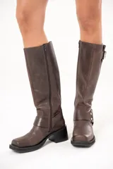 Bota biker de caña alta color marrón, confeccionada en cuero. Presenta hebilla en la parte superior, detalle de aro en la parte inferior y cierre lateral.