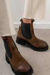 Bota tipo Chelsea de cuero marrón con suela track negra de goma. Presenta paneles elásticos laterales y una costura reforzada en la puntera. Incluye una correa de cuero desmontable con hebilla metálica.