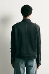 Chaqueta cropped fit de denim de algodón en color negro con efecto lavado, cuello mao y cierre frontal con cremallera. Presenta aplicaciones de trabillas horizontales en el pecho y en las mangas, además de hombreras abotonadas.