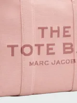 Bolso tipo tote bag de Marc Jacobs en color beige, confeccionado en lona resistente. Presenta el texto "THE TOTE BAG" y "MARC JACOBS" impreso en la parte frontal en color negro. Cuenta con asas superiores y una correa de hombro ajustable y extraíble, además de ribetes laterales negros con inscripciones.