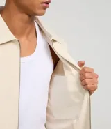 Campera masculina de corte boxy en color beige, con cuello inglés, cierre frontal de cremallera, mangas largas con puños elásticos y ruedo con elástico. Presenta bolsillos laterales y detalles de recortes en el diseño.
