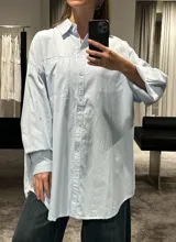 Camisa de corte oversize color marrón, con cuello clásico, cierre frontal de botones y mangas largas con puños ajustables. Presenta un diseño de hombros caídos y un bolsillo frontal en el pecho.