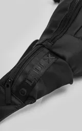 Riñonera negra confeccionada en cordura impermeable de alta resistencia. Presenta tres compartimentos principales con cierres metálicos, un bolsillo lateral adicional y una correa ajustable de alta densidad con broche de seguridad. Incluye una chapa metálica decorativa en el frente.