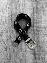 Cinto negro de charol con hebilla metálica plateada con diseño labrado y ojales metálicos.