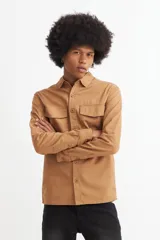 Camisa marrón de manga larga con cuello clásico, cierre frontal con botones, dos bolsillos con solapa abotonada en el pecho y puños abotonados.