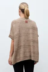 Capa tejida de punto en color beige, con diseño abierto y cierre frontal mediante broche metálico con detalles en cuero sintético negro. Presenta mangas cortas y textura suave.