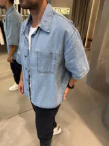 Camisa de jean de corte boxy, color azul claro, con mangas cortas, cuello clásico y cierre frontal con botones metálicos. Presenta un bolsillo frontal en el pecho.