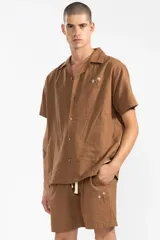 Camisa de hombre color marrón claro, de calce relajado y corte cuadrado, confeccionada en lino y algodón. Tiene cuello tipo camp, cierre con botones de coco grabados con el logo y bordado sutil de flores en el pecho izquierdo.