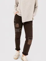 Pantalón de corte relajado color marrón oscuro, con parches decorativos de lentejuelas y tachas metálicas. Presenta cintura elástica con cordón ajustable y texto estampado en una de las piernas.