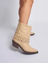 Bota de cuero color marrón con caña media, taco cuadrado y punta fina. Presenta tachas plateadas en el borde superior y en la tira que rodea el tobillo. El cuero tiene un acabado liso y brillante.
