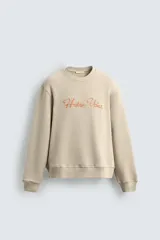 Sudadera de algodón color beige claro, corte relajado, cuello redondo y manga larga. Presenta bordado en relieve en el frente con texto en cursiva de color cobre.