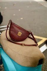 Cartera de mano pequeña con diseño semicircular, confeccionada en cuero con textura tipo croco color marrón. Presenta una placa metálica dorada con las iniciales AL en el centro y herrajes de bronce que permiten ajustar la correa de diversas formas.