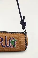Bolso tipo bandolera de formato cilíndrico, confeccionado con material trenzado en color tostado, con detalle de bordado frontal que dice 'Rio' acompañado de una palmera y un tucán. Cuenta con una correa larga de cuerda oscura con nudos ajustables y cierre superior.