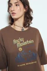Remera blanca de manga corta con calce relajado y cuello a la base. Presenta un estampado frontal pequeño y una estampa protagonista en la espalda con el texto "Rocky Mountain National Park" y un paisaje de montañas y árboles.