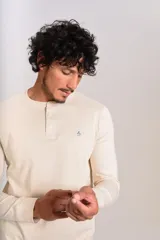 Remera de manga larga tipo Henley, color beige, con cuello redondo y tapeta con botones. Presenta un pequeño logo bordado en el pecho.