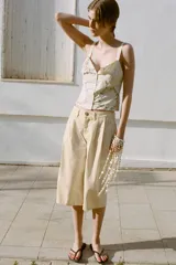 Top de color crema con escote en pico, tirantes anchos y cierre frontal de botones. Presenta bordados florales en tono verde claro alrededor del escote y en forma de semicírculo en la parte inferior delantera.