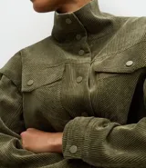 Campera de pana cotelé color verde militar, con cuello camisero, cierre frontal con botones metálicos y bolsillos frontales con solapa. Presenta diseño de sisa desplazada y cordones ajustables en el ruedo.
