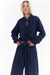 Blusa de denim de algodón super liviano, de estilo oriental en color azul con costuras a contratono rojas. Presenta cierre frontal con lazos tipo moño, cuello mao y mangas largas abullonadas con puños elásticos.