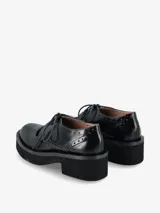 Zapato tipo guillermina con cordones, confeccionado en cuero negro con detalles de perforaciones estilo brogue. Presenta una suela de plataforma gruesa y tacón cuadrado.