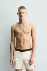 Short de baño blanco marfil con cintura marrón y cordón ajustable del mismo color. Presenta franjas laterales en tono azul marino con finos bordes rojizos y bolsillos laterales.