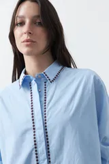 Camisa blanca de manga larga con detalles bordados en contraste a lo largo de la botonadura, el cuello y los puños. Presenta una silueta relajada y un diseño clásico con un toque artesanal.