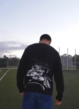 Buzo de punto acanalado en color negro, con diseño gráfico estampado en blanco en la espalda que representa una vista aérea de un estadio. Presenta mangas largas y un corte holgado.