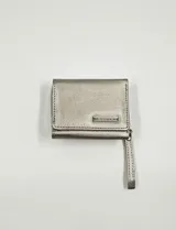 Billetera pequeña de diseño compacto en color plateado metalizado, con textura granulada y cierre de solapa. Incluye una placa metálica con el logo de la marca y una correa de mano lateral.