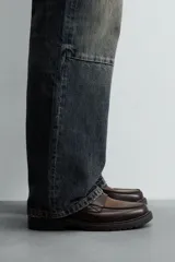 Mocasín de cuero marrón oscuro con antifaz en tono camel. Presenta horma redonda y suela track gruesa de goma negra.