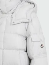 Campera puffer Moncler Maya color gris claro, rellena de plumas, con capucha desmontable, cierre frontal con botones a presión y bolsillos laterales con cierre.