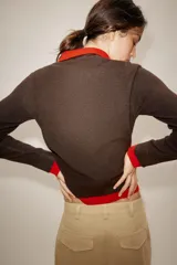 Jersey de punto con efecto doble, cuello subido y manga larga. Presenta un diseño en color marrón con detalles en rojo vibrante en el cuello, puños y ruedo. Cuenta con cierre frontal parcial mediante botones.
