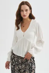 Blusa blanca de viscosa, con cuello en V, detalles de frunces en el frente, botones decorativos y mangas largas holgadas.