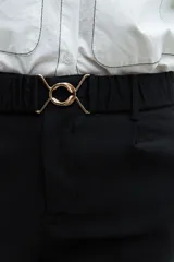 Pantalón negro de vestir, corte recto y tiro alto, con puños doblados. Se ajusta a la cintura con un cinturón fino de hebilla metálica con forma de doble 'X' o infinito.