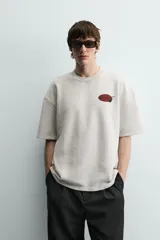 Sudadera gris de corte relaxed fit, cuello redondo y manga corta. Presenta un estampado en el pecho con la frase "Tokyo Kitchen" en un óvalo rojo con palillos.