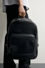 Mochila de acabado liso en color negro, con compartimento principal con cierre de cremallera, bolsillo frontal, detalle de hebillas en los laterales y asas acolchadas.