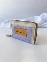 Billetera pequeña con cierre metálico, cuerpo principal de cuero lila texturizado y ribetes en color crema. Presenta un parche de cuero marrón con la palabra "chowie" escrita en cursiva.