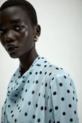 Camisa de manga larga con cuello fluido y diseño cruzado en el frente. Confeccionada en tejido semitransparente de color celeste con estampado de lunares negros. Presenta bajo asimétrico y cierre en la espalda con botones forrados.