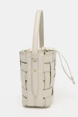 Bolso tipo bucket de diseño trenzado en color marrón oscuro con costuras a la vista en contraste. Cuenta con asa de mano fija, correa bandolera extraíble y compartimento interior de tela en color crudo con cierre mediante fruncido.