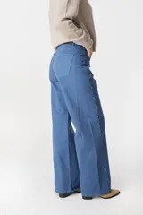 Jean de corte recto y tiro medio, color azul, con detalles de tachas metálicas doradas a lo largo de las costuras laterales externas.