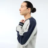 Buzo de felpa gris con cuello en V, detalles en azul marino y ribetes rayados en el cuello, puños y ruedo. Tiene corte holgado y mangas largas con paneles en contraste.