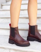 Botas tipo Chelsea de cuero color marrón, con paneles laterales elásticos en negro y tirador trasero en color rojo. Presentan una suela track robusta de goma negra con costuras decorativas en rojo.