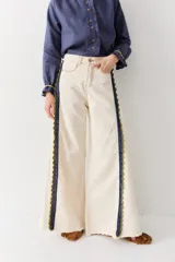 Jean de corte wide leg en color crudo, con detalle de franjas laterales tejidas en crochet azul y verde. Presenta costuras en contraste y ruedo deshilachado.