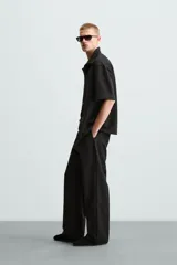 Sobrecamisa negra de tejido con elasticidad, cuello solapa y manga corta. Tiene dos bolsillos de plastrón en el pecho y cierre frontal con botones a presión.