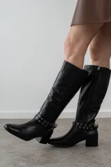 Bota alta de cuero negro, con punta fina y taco cuadrado. Presenta una correa con ojales metálicos alrededor del tobillo y cierre lateral.
