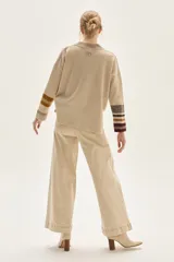 Sweater tejido de lambswool color beige, con cuello redondo y puños acampanados con rayas de colores (marrón, mostaza, bordó y beige). Incluye un detalle de temporada hecho a mano en la espalda.