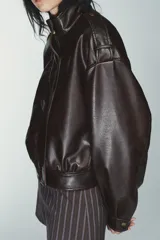 Cazadora corta de efecto piel color marrón oscuro, con cuello subido y cierre frontal oculto con cremallera y botones a presión. Presenta mangas largas con puños y pliegues, trabillas en los hombros, bolsillos laterales y bajo abullonado. El tejido tiene un efecto lavado.